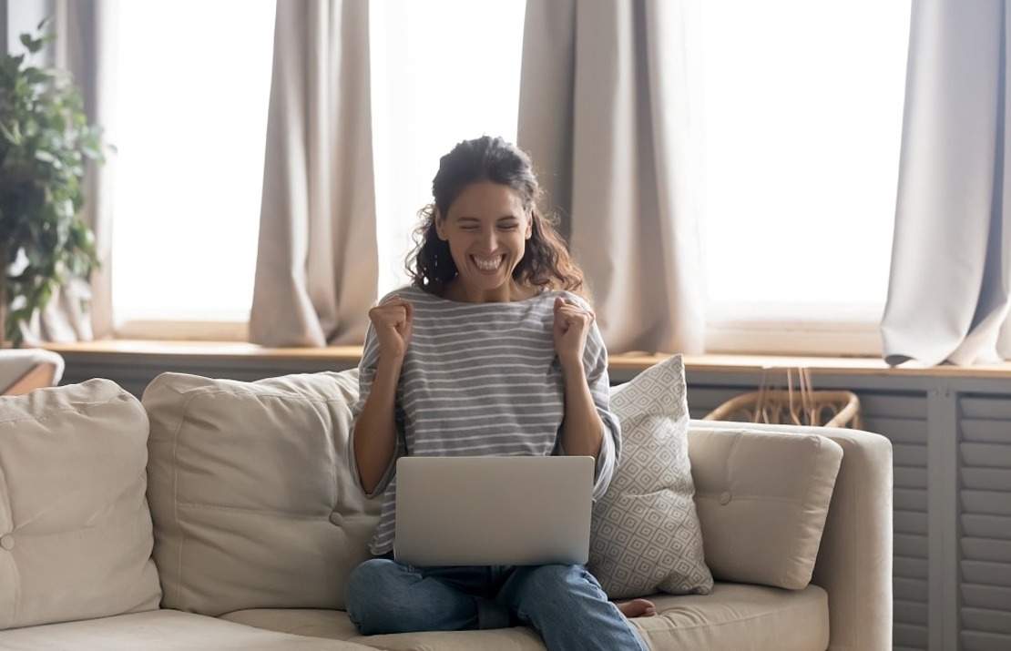 Eine Frau sitzt mit einem Laptop auf dem Schoß auf einem Sofa, lächelt und reckt aufgeregt die Fäuste in die Höhe. Im Hintergrund fällt Sonnenlicht durch große Fenster, an der Seite ist eine Topfpflanze zu sehen.