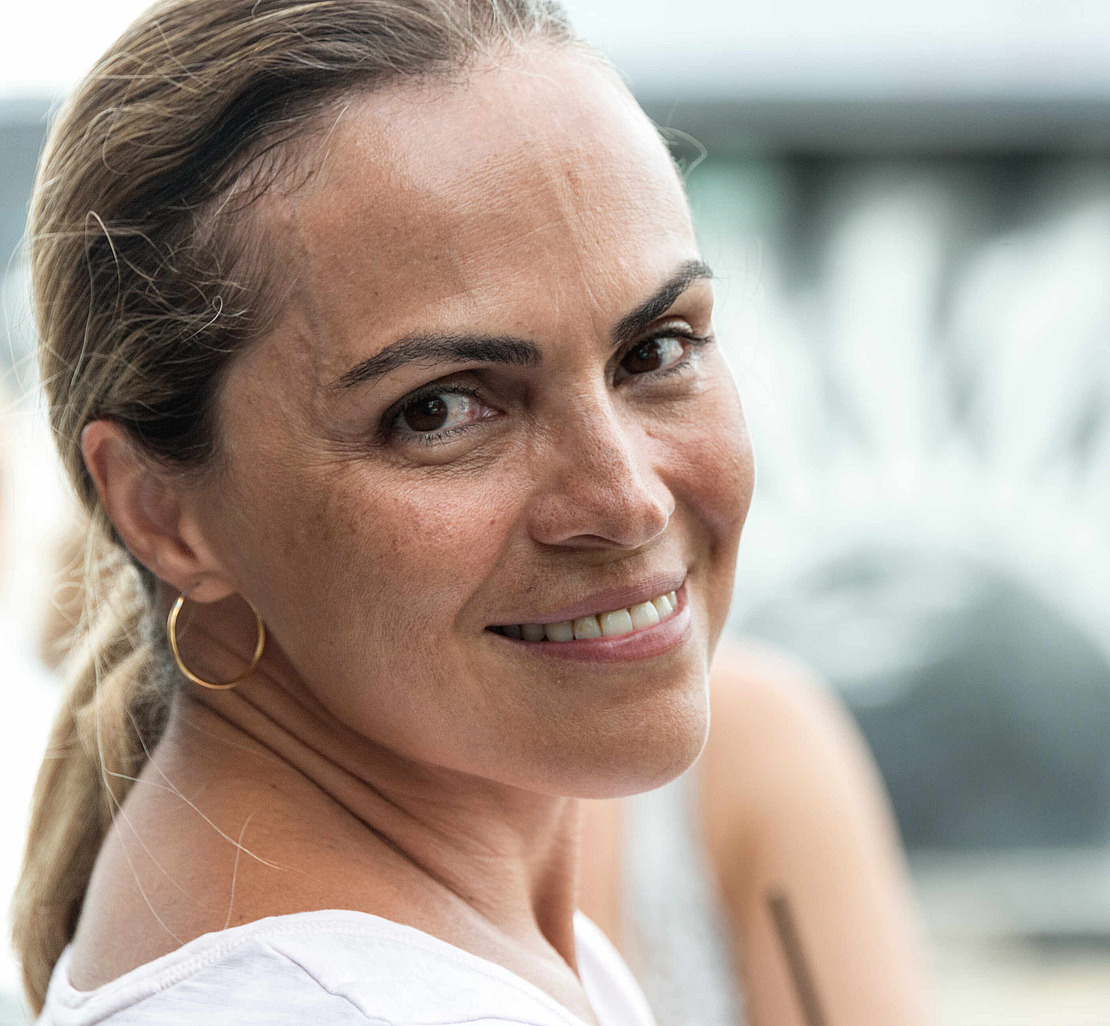 Eine Frau mit zurückgekämmtem hellbraunem Haar lächelt in die Kamera. Sie trägt ein weißes Oberteil und goldene Creolen. Der Hintergrund ist unscharf und zeigt eine weitere Person sowie ein kreisförmiges schwarz-weißes Muster.