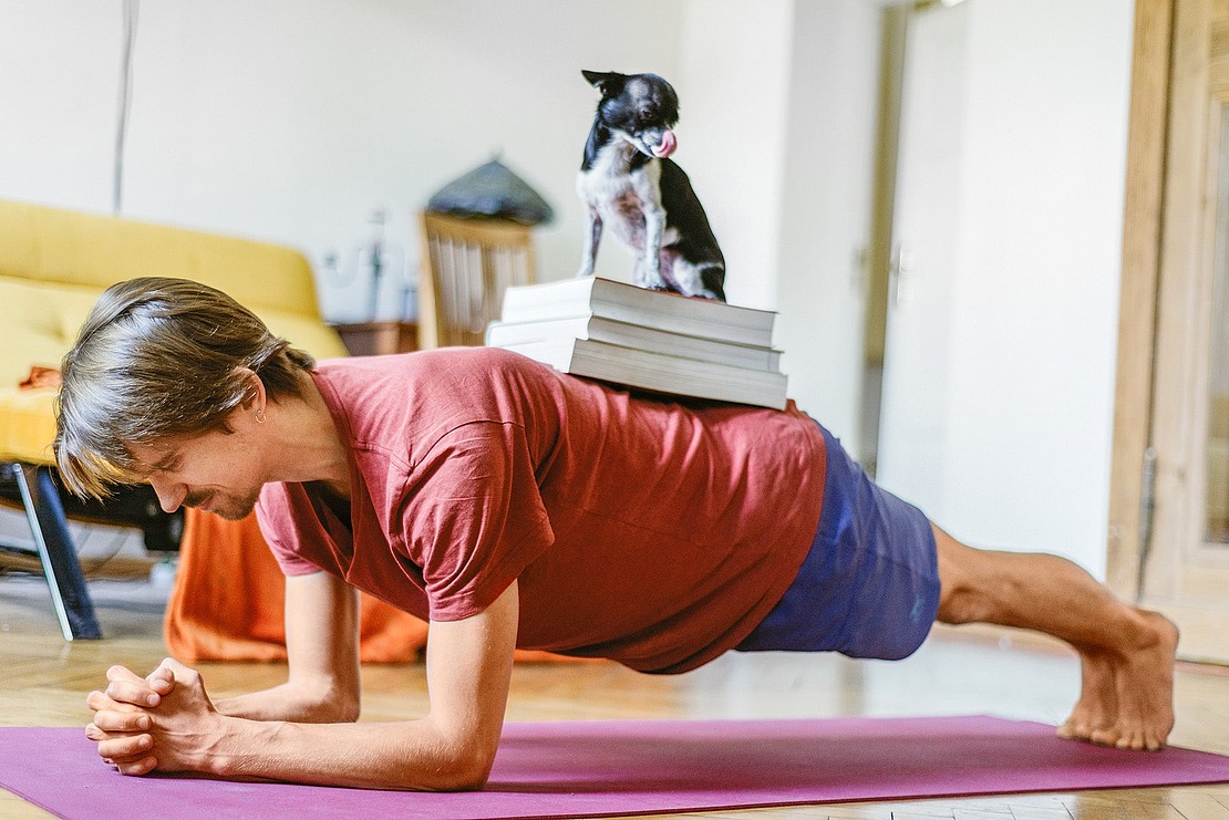Ein Mann macht eine Planke auf einer Yogamatte in einem Raum und balanciert dabei mehrere Bücher und einen kleinen Hund auf seinem Rücken.