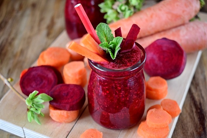 Ein erfrischendes Glas Rote-Bete- und Karottensaft, garniert mit Minzblättern, umgeben von geschnittenen Rüben und Karotten auf einem Holzbrett. Im Hintergrund liegen zwei ganze Karotten.