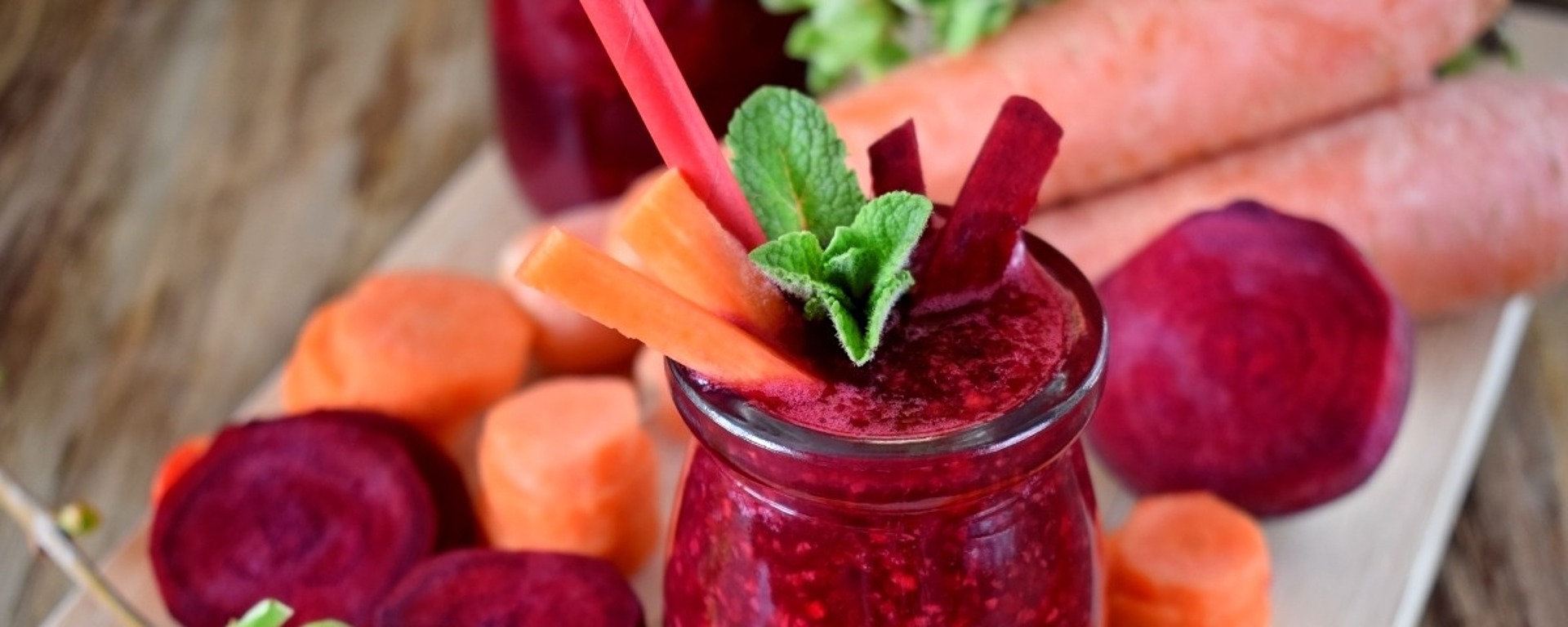 Ein erfrischendes Glas Rote-Bete- und Karottensaft, garniert mit Minzblättern, umgeben von geschnittenen Rüben und Karotten auf einem Holzbrett. Im Hintergrund liegen zwei ganze Karotten.