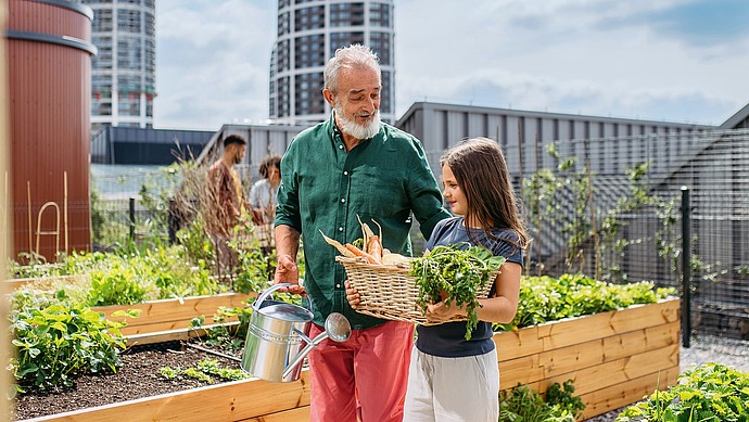 Ein älterer Mann und ein junges Mädchen stehen in einem städtischen Garten. Der Mann hält eine Gießkanne, während das Mädchen einen Korb mit frischem Gemüse trägt. Im Hintergrund erheben sich moderne Gebäude unter einem teilweise bewölkten Himmel.