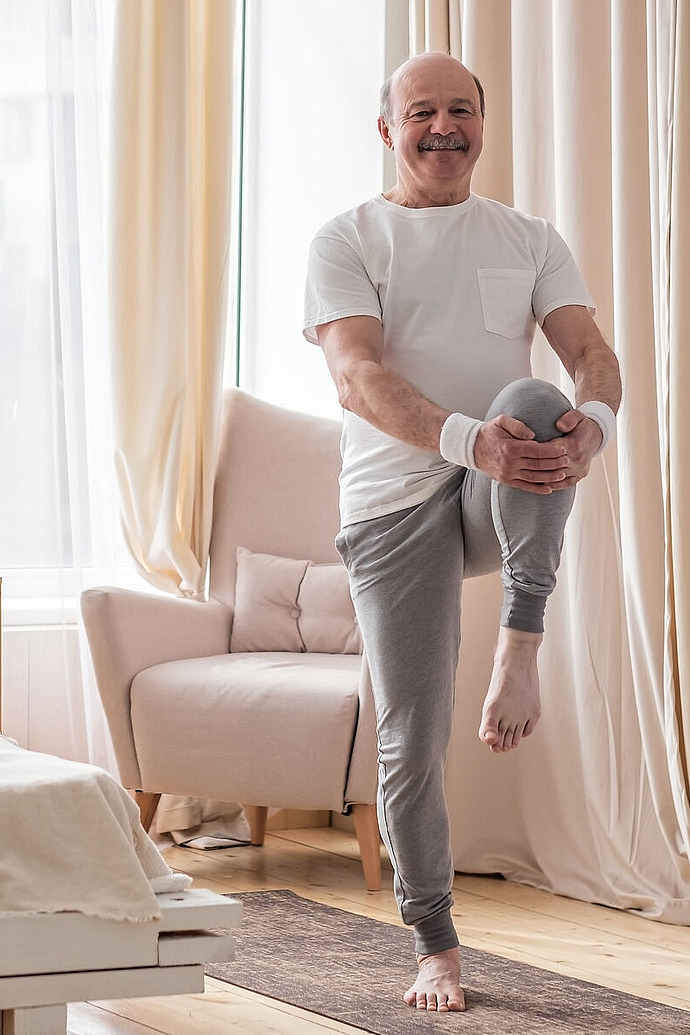 Ein älterer Mann in Sportkleidung balanciert auf einem Bein und zieht dabei sein Knie an die Brust. Er befindet sich in einem hellen, gemütlichen Raum mit lichtdurchlässigen Vorhängen, einem Sessel und einem Bett, das im Hintergrund zu sehen ist.
