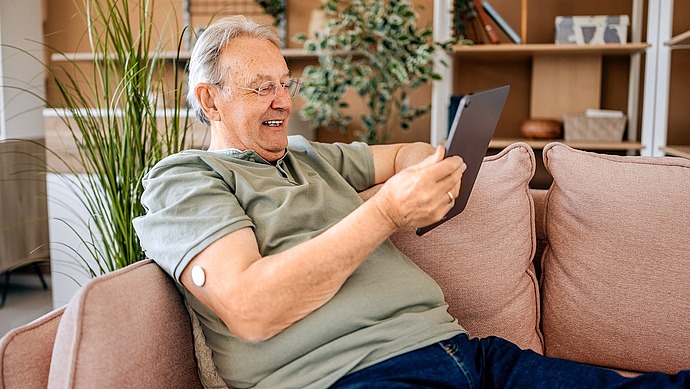Ein lächelnder älterer Mann mit Brille sitzt auf einem Sofa und hält ein Tablet in der Hand. Er trägt ein grünes Hemd und hat ein medizinisches Pflaster am Oberarm. Im Hintergrund sind Regale, Pflanzen und gemütliche Dekoration zu sehen.