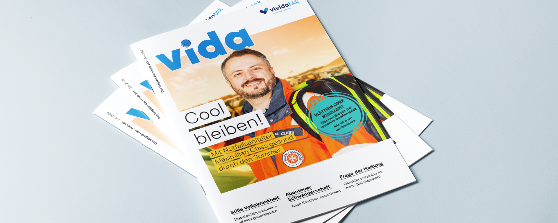 Auf einer hellblauen Fläche liegt ein Stapel „Vida“-Magazine, auf dem oberen Cover ist ein lächelnder Mann in einer Warnweste zu sehen, mit der Überschrift „Cool bleiben!“ und einem Text zur Sommersicherheit für Arbeitnehmer.
