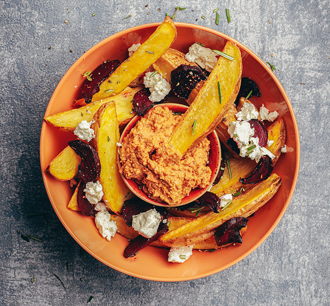 Oranger Teller mit gerösteten Kartoffelspalten, Rübenscheiben, Ziegenkäse und einer kleinen Schale mit orange-rotem Dip in der Mitte. Der Teller ist mit Kräutern auf einer grau strukturierten Oberfläche garniert.