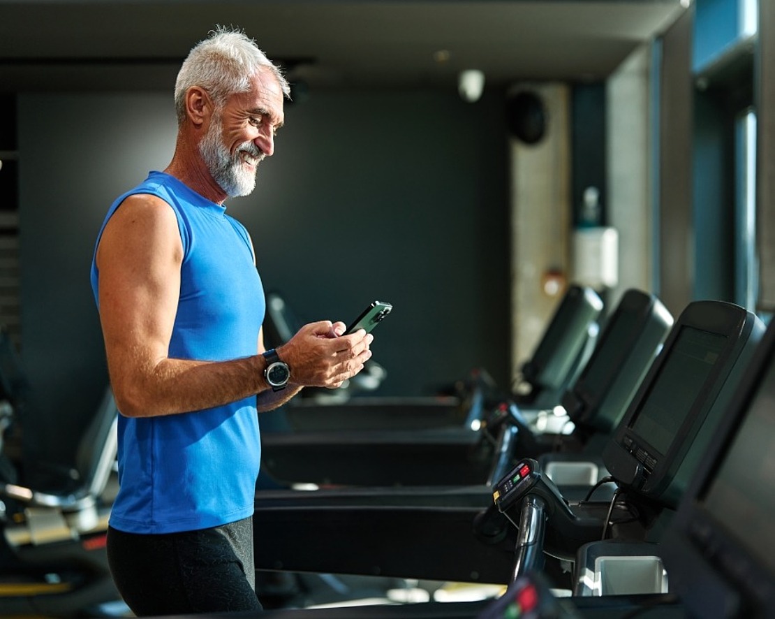 Ein älterer Mann mit weißem Bart und kurzen Haaren, der ein blaues ärmelloses Hemd trägt, steht auf einem Laufband in einem Fitnessstudio. Er lächelt und schaut dabei auf sein Smartphone. Im Hintergrund sind mehrere Laufbänder und ein Fenster zu sehen.