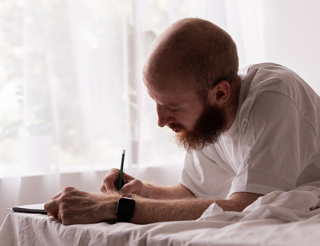 Ein Mann mit Bart und weißem T-Shirt liegt in einem hellen Zimmer auf einem Bett. Er schreibt mit einem Stift in ein Notizbuch und trägt eine Smartwatch. Weiches Licht fällt durch die transparenten Vorhänge hinter ihm.