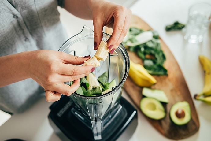 Eine Person gibt eine Banane in einen Mixer mit Blattgemüse. Auf einem Holzschneidebrett daneben liegen geschnittene Avocado, eine ganze Banane und weiteres Blattgemüse. Die Szene lässt auf die Zubereitung eines gesunden Smoothies schließen.