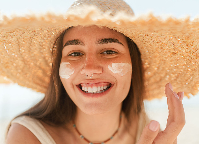 Eine lächelnde Frau mit großem Strohhut trägt am Strand Sonnencreme auf ihre Wangen auf. Sie hat langes braunes Haar, einen Nasenring und blickt an einem sonnigen Tag direkt in die Kamera.