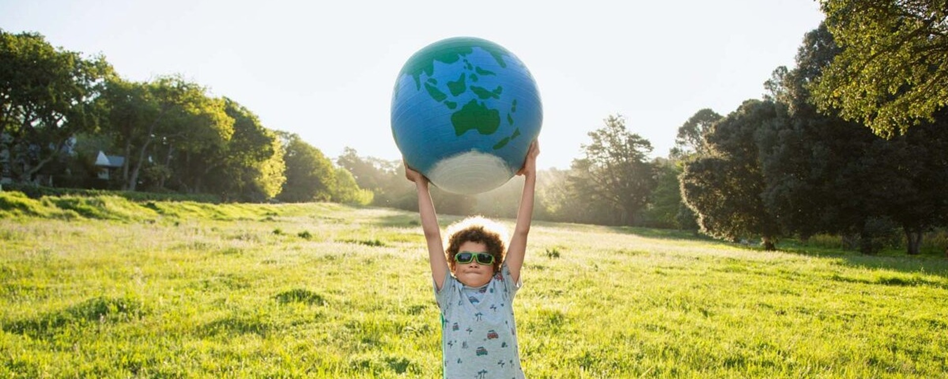 Ein Junge mit Sonnenbrille und Sommerkleidung steht auf einer sonnigen Wiese und hält eine große aufblasbare Weltkugel über seinen Kopf, während im Hintergrund Bäume und Grünflächen zu sehen sind.