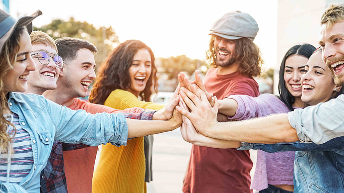 Eine Gruppe junger Erwachsener steht im Freien in einem Kreis, lächelt und legt ihre Hände in der Mitte zusammen, um Einigkeit und Freundschaft zu zeigen. Der Hintergrund ist sonnenbeschienen und alle sehen glücklich und engagiert aus.