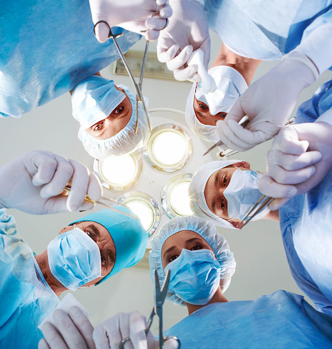 OP-Ärzte mit Masken und Arbeitskleidung beugen sich mit medizinischen Instrumenten nach unten und schauen in die Kamera