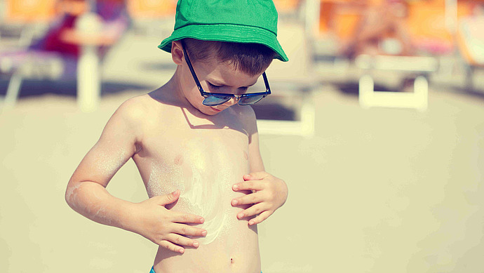 Ein Junge mit grünem Hut und Sonnenbrille trägt Sonnencreme auf seine Brust auf, während er an einem sonnigen Strand steht. Der Hintergrund ist unscharf und zeigt Liegestühle und andere Strandbesucher.