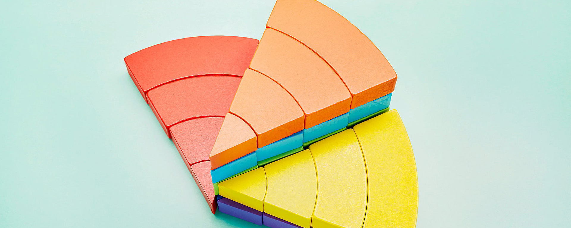 Bunte Halbkreisblöcke aus Holz in Rot-, Orange- und Gelbtönen sind fächerförmig auf einem hellblauen Hintergrund angeordnet. Die Teile passen wie Puzzle-Scheiben zusammen und bilden ein geometrisches Muster.
