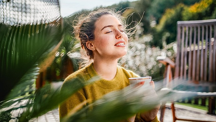 Eine Frau sitzt im Freien mit geschlossenen Augen, lächelt sanft und hält eine Tasse. Das Sonnenlicht beleuchtet ihr Gesicht, und das grüne Laub im Vorder- und Hintergrund schafft eine friedliche und entspannte Atmosphäre.