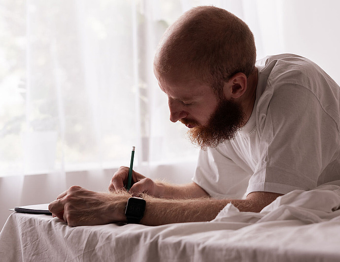 Ein Mann mit Bart und weißem T-Shirt liegt auf einem Bett und schreibt in ein Notizbuch. Sonnenlicht fällt durch die transparenten Vorhänge im Hintergrund und schafft eine ruhige, helle Atmosphäre.