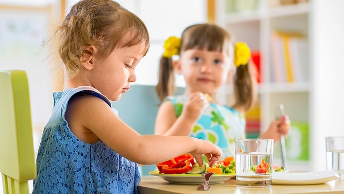 Zwei kleine Mädchen sitzen an einem Tisch und essen. Ein Mädchen in einem blauen Kleid ist darauf konzentriert, geschnittene rote Paprika von ihrem Teller zu nehmen, während das andere Mädchen mit gelben Haargummis zuschaut. Auf dem Tisch stehen Gläser mit Wasser.