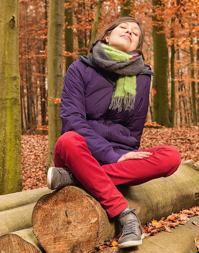 Eine Person, die einen Schal, eine Jacke und eine rote Hose trägt, sitzt im Schneidersitz auf einem Baumstamm in einem Wald mit Herbstlaub, hat die Augen geschlossen, lächelt friedlich und genießt die freie Natur.