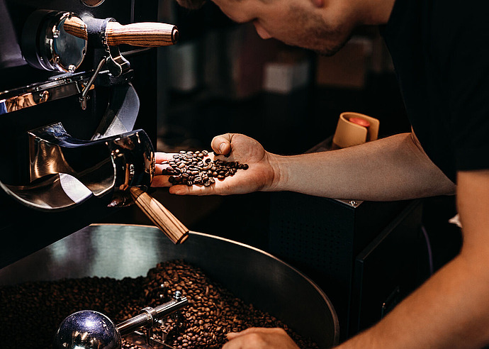 Eine Person untersucht geröstete Kaffeebohnen in ihrer Hand neben einer großen, mit Bohnen gefüllten Kaffeeröstmaschine in einer Kaffeerösterei.