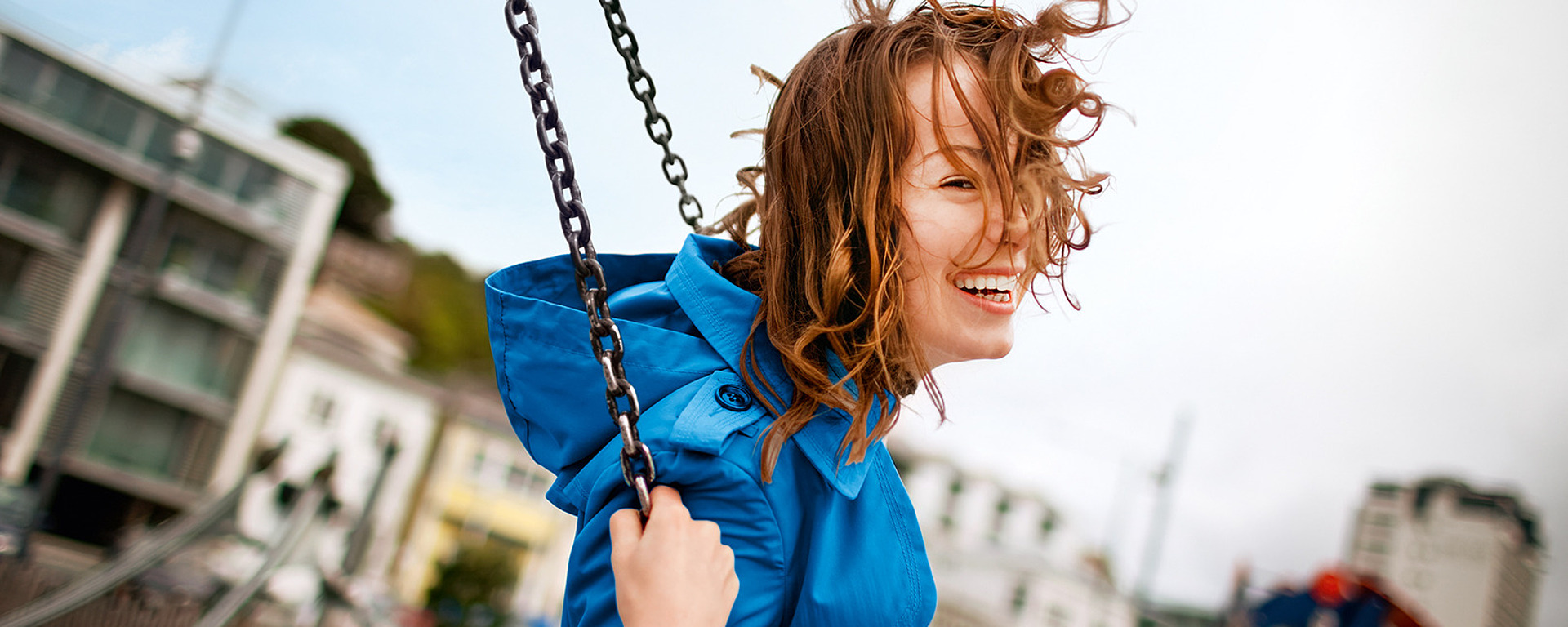 Eine Person in einem blauen Mantel lächelt freudig, während sie auf einem Spielplatz schaukelt, wobei ihr Haar im Wind weht. Im Hintergrund sind unscharfe Gebäude und Spielgeräte zu sehen.