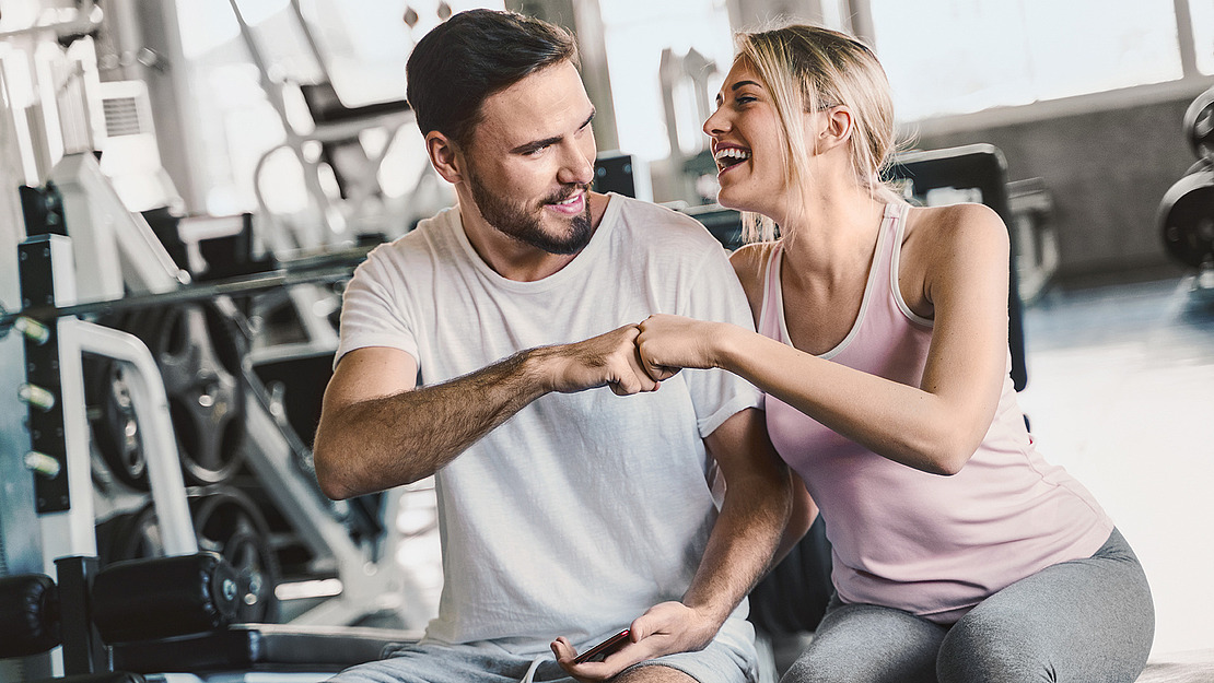 Das Bild zeigt einen Mann und eine Frau im Fitnessstudio, die gemeinsam auf ein Smartphone schauen.