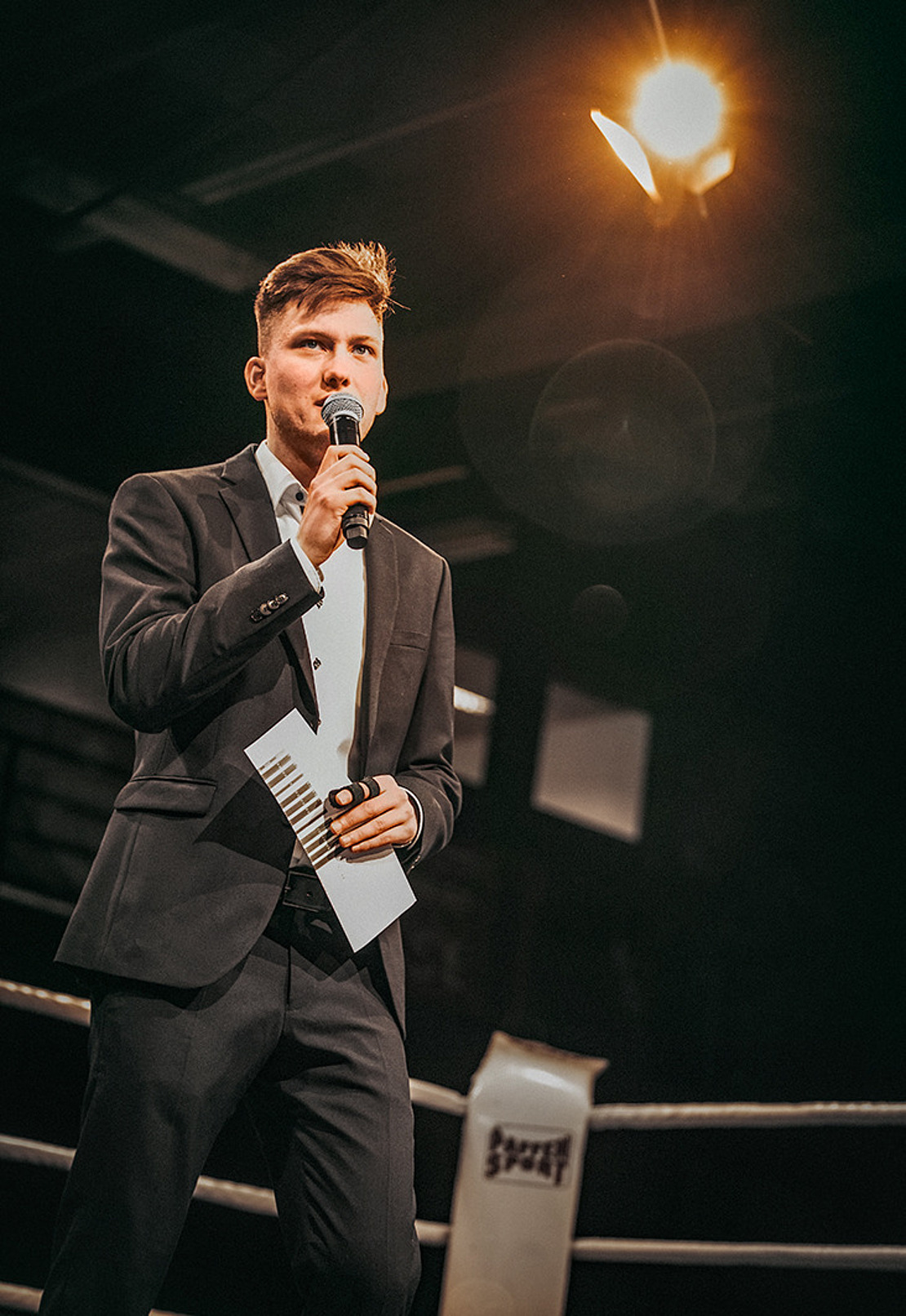 Ein Mann im Anzug steht in einem Boxring, hält ein Mikrofon und Notizkarten in der Hand und spricht unter einem hellen Scheinwerferlicht. Der Hintergrund ist dunkel und unterstreicht die dramatische Kulisse.