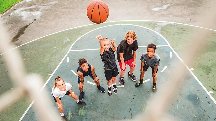Fünf Kinder auf einem Basketballplatz im Freien blicken auf, als einer von ihnen einen Basketball in Richtung Korb wirft, und alle scheinen begeistert und engagiert zu spielen.