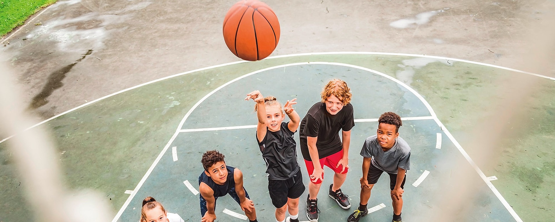 Fünf Kinder auf einem Basketballplatz im Freien blicken auf, als einer von ihnen einen Basketball in Richtung Korb wirft, und alle scheinen begeistert und engagiert zu spielen.