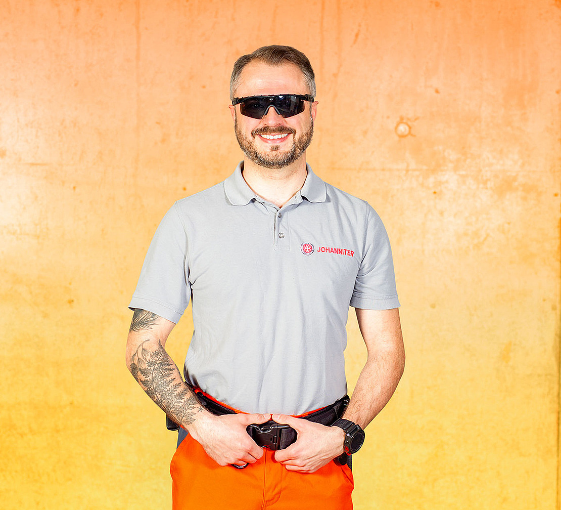 Ein Mann mit Bart, Sonnenbrille und einem Tattoo auf dem linken Arm steht lächelnd vor einer orangefarbenen Wand. Er trägt ein hellgraues Poloshirt, eine orangefarbene Hose und eine schwarze Uhr. Seine Hände ruhen auf seinem Gürtel.