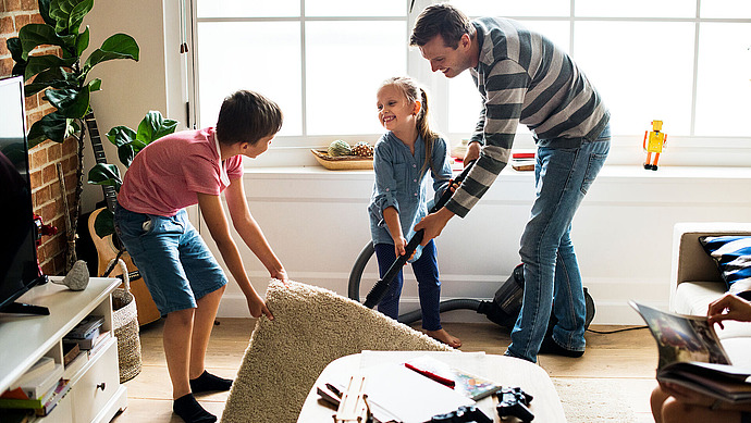 Ein Vater und zwei Kinder saugen und räumen ein helles Wohnzimmer auf. Vater und Tochter bedienen den Staubsauger, während der Sohn einen kleinen Teppich hochhebt. Der Raum ist mit Pflanzen und Spielzeug dekoriert, Sonnenlicht fällt durch das Fenster.