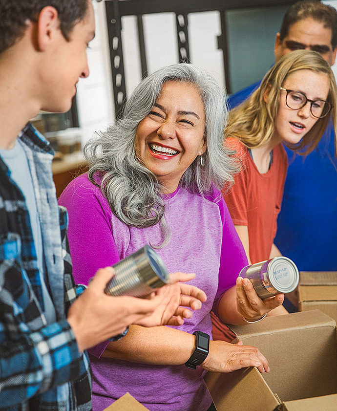 Eine Gruppe von Menschen packt fröhlich Konserven in Kartons. Wahrscheinlich sind sie ehrenamtlich tätig oder helfen bei einer Lebensmittelsammlung. Eine Frau mit grauem Haar in einem lila Hemd lächelt und hält eine Dose in der Hand.
