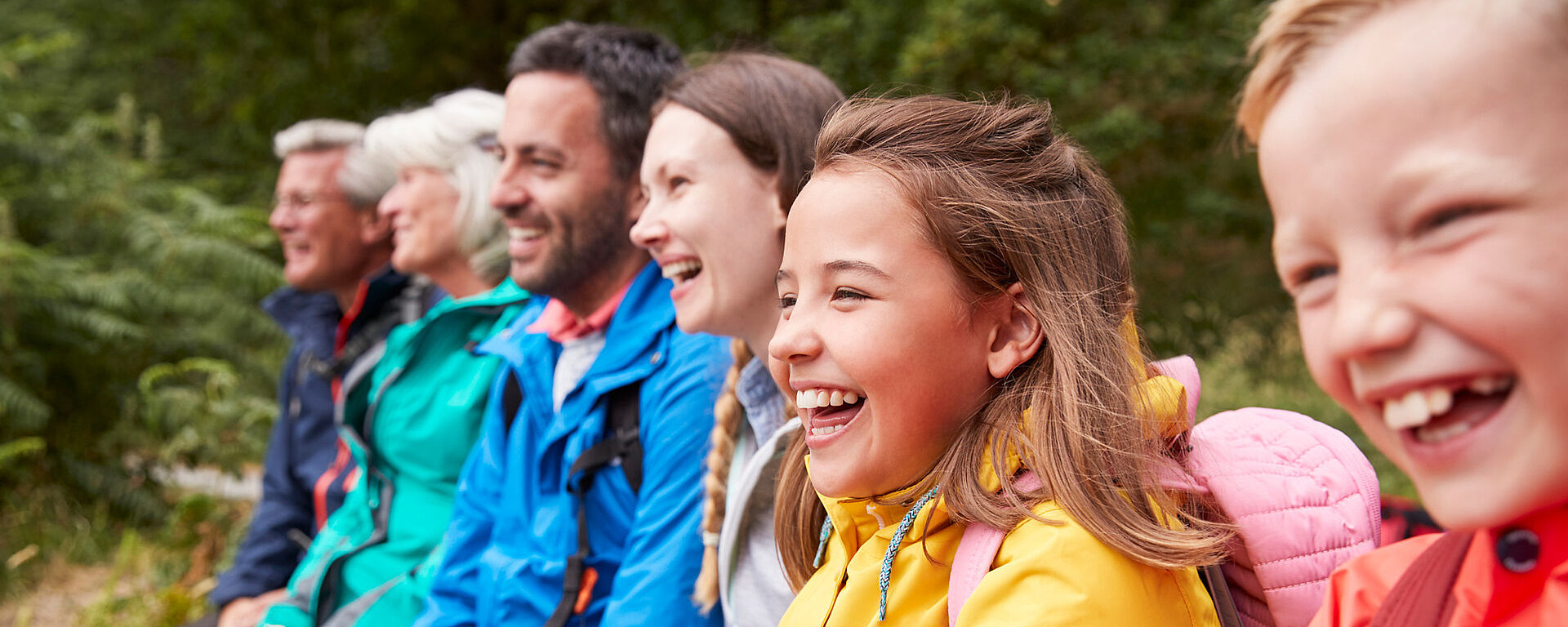 Eine generationsübergreifende Gruppe, darunter Kinder und Erwachsene, sitzt in bunten Jacken im Freien, lächelt und lacht zusammen, während im Hintergrund Grünflächen zu sehen sind.