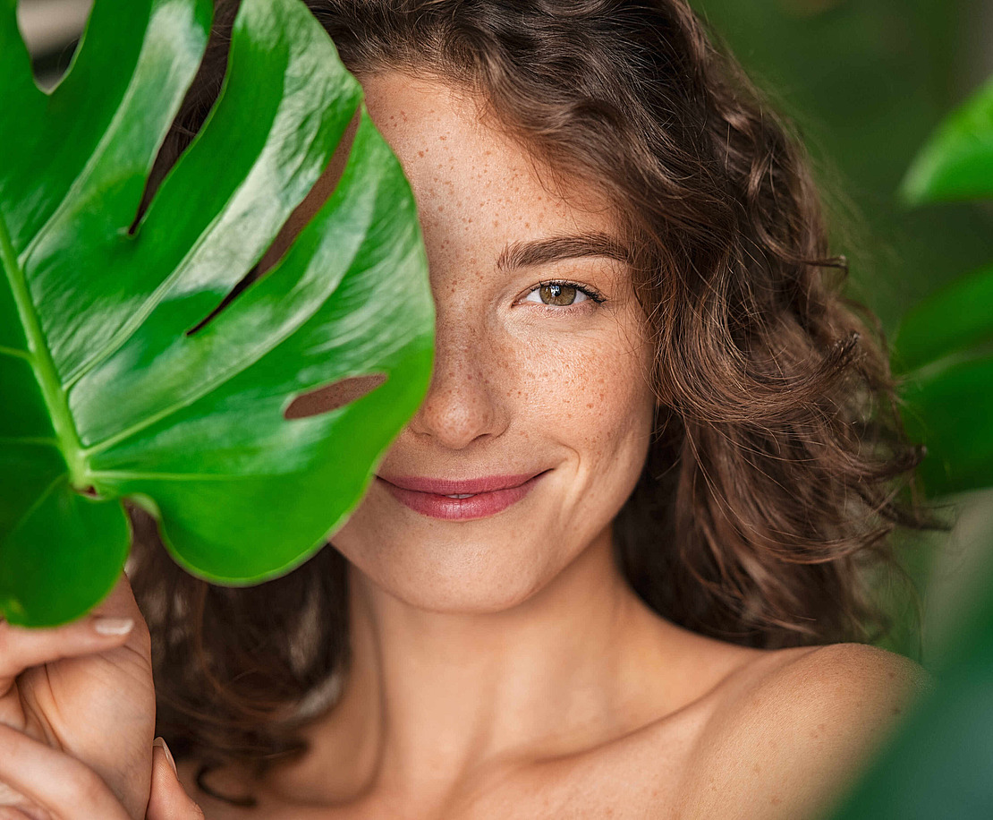 Lächelnde Frau mit lockigem Haar und Sommersprossen hält ein großes grünes Monsterablatt, das eine Seite ihres Gesichts teilweise bedeckt, mit üppigem grünem Laub im Hintergrund.