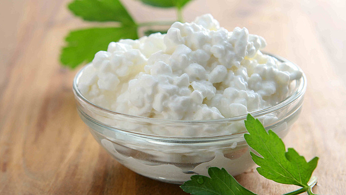 Eine mit Hüttenkäse gefüllte Glasschale steht auf einer Holzplatte. Frische grüne Petersilienblätter werden zur Dekoration um die Schale herum und teilweise darunter gelegt.