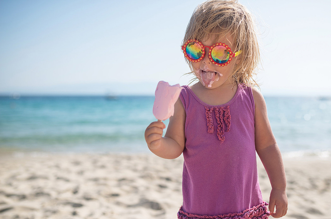 Ein Kind mit Sonnenbrille isst ein Eis am Strand.