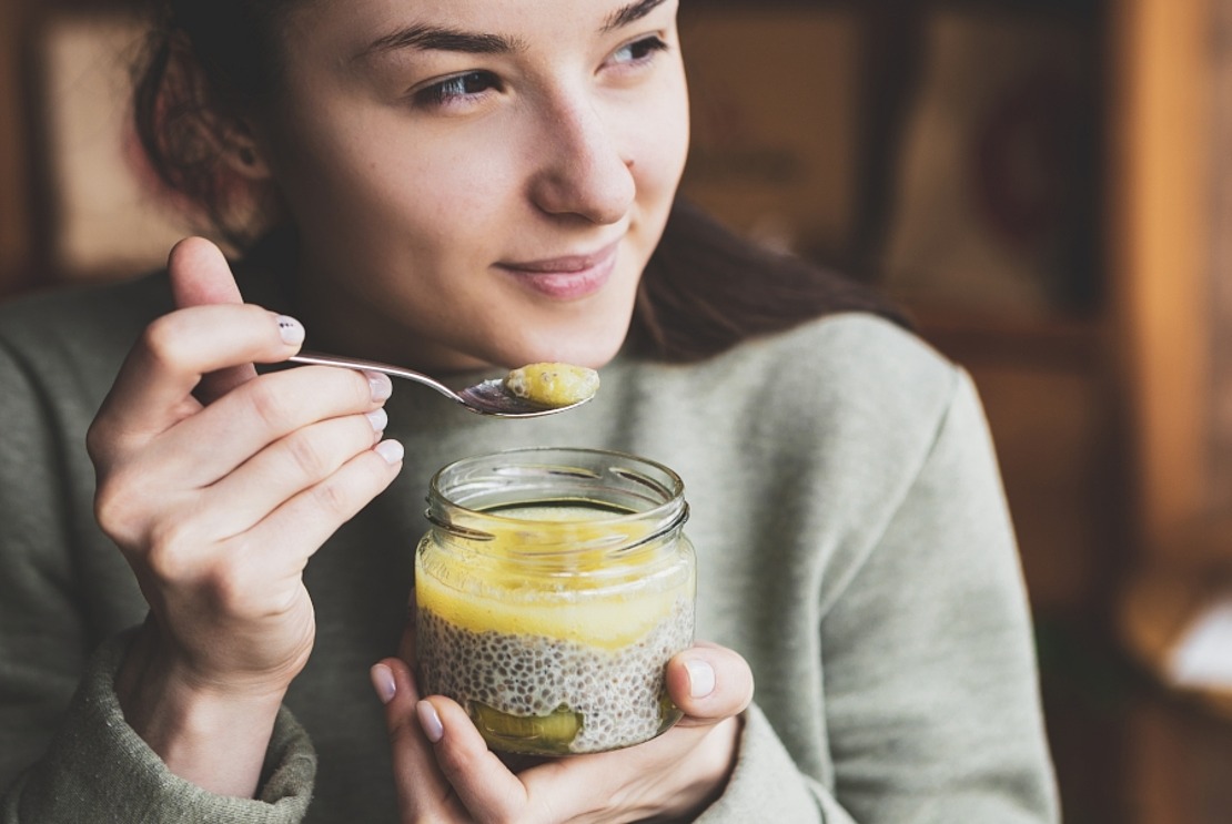 Eine Person im grünen Pullover hält ein Glas Chia-Pudding mit gelbem Topping, lächelt leicht und hält einen Löffel des Desserts an den Mund. 