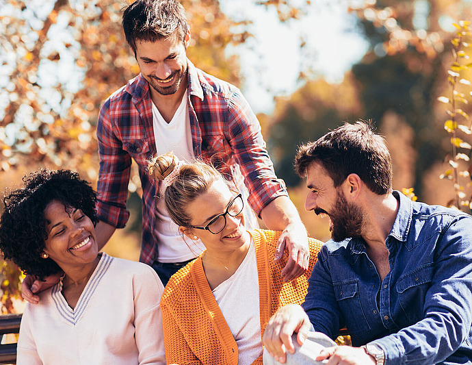 Vier Freunde sitzen und stehen im Herbst zusammen auf einer Bank im Freien, lächeln und lachen zwischen goldenem Laub. Alle wirken glücklich und genießen die Gesellschaft des anderen.