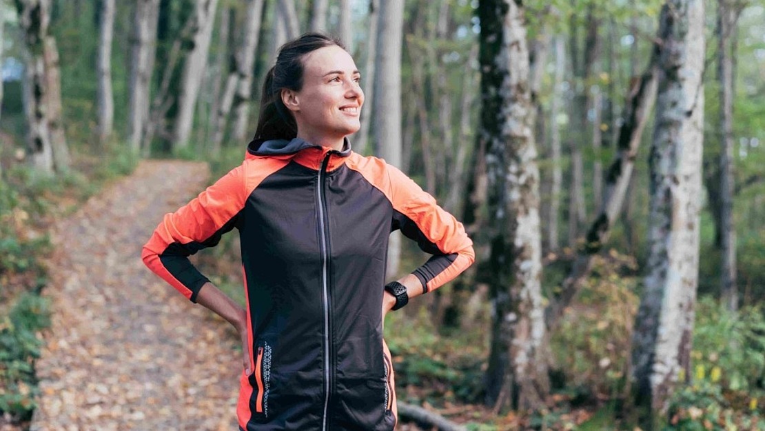 Eine Frau in einer schwarz-orangenen Sportjacke steht auf einem Waldweg, lächelt und schaut mit den Händen in den Hüften zur Seite. Der Weg ist mit abgefallenen Blättern bedeckt und von hohen Bäumen umgeben.