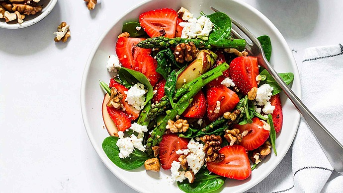 Eine Schüssel Spinatsalat mit geschnittenen Erdbeeren, geröstetem Spargel, zerbröckeltem Feta und Walnüssen, daneben eine Gabel. Daneben stehen eine kleine Schale mit Walnüssen und ein Glas Honig auf einer Marmorplatte.