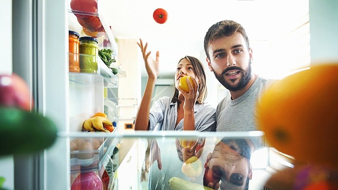 Ein Mann und eine Frau schauen in einen offenen Kühlschrank. Die Frau wirft eine Tomate in die Luft, während sie einen Apfel hält. Im Kühlschrank sind verschiedene Früchte, Gemüse und Gläser zu sehen.