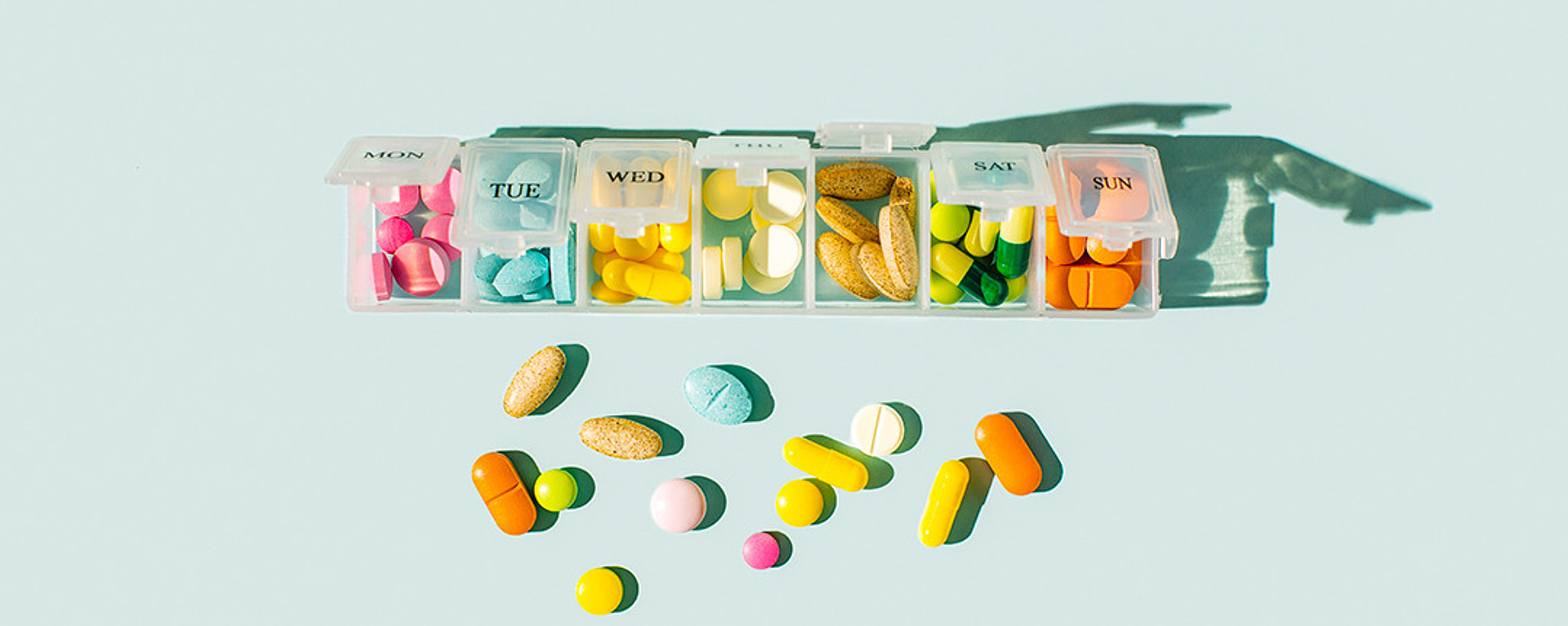 Ein wöchentlicher Pillen-Organizer, bei dem jedes Fach offen ist und verschiedene bunte Pillen und Tabletten zu sehen sind. Weitere Pillen sind vor einem türkisfarbenen Hintergrund verstreut. Schatten werden hinter den Pillen-Organizer geworfen.