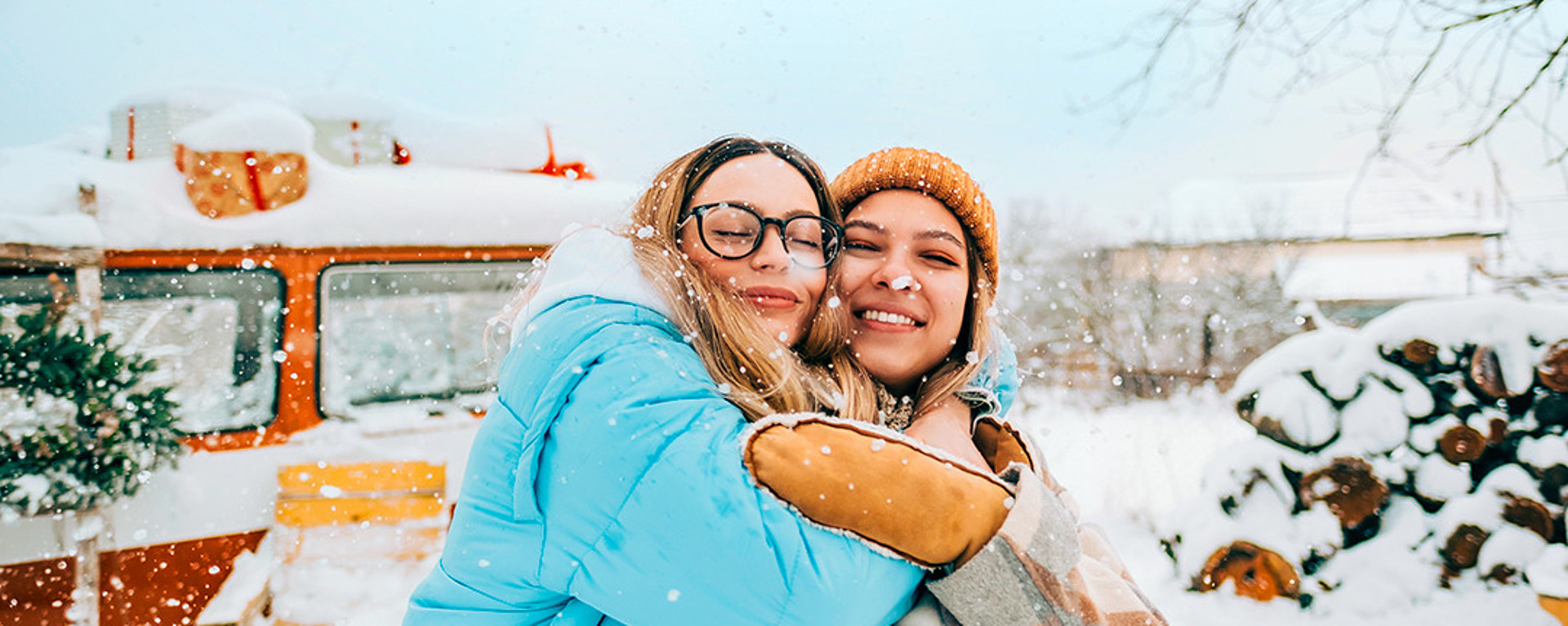 Zwei Freunde lächeln und umarmen sich in einer verschneiten Umgebung. Einer trägt eine blaue Pufferjacke und eine Brille, der andere einen karierten Mantel und eine Strickmütze. Im Hintergrund sind ein Oldtimer-Wohnmobil und ein Stapel Brennholz zu sehen, um sie herum fällt sanft Schnee.