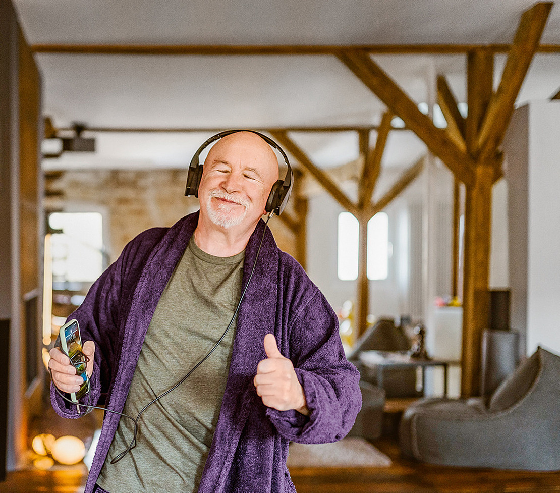 Ein älterer Mann in einem lilafarbenen Bademantel und mit Kopfhörern lächelt und zeigt den Daumen nach oben, während er ein Telefon in der Hand hält.