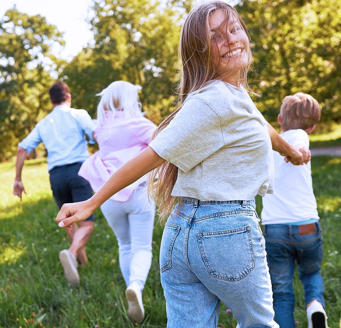 Ein lächelndes Mädchen blickt zurück, während es an den Händen haltend mit seiner Familie durch einen sonnigen, grasbewachsenen Park mit grünen Bäumen im Hintergrund läuft.