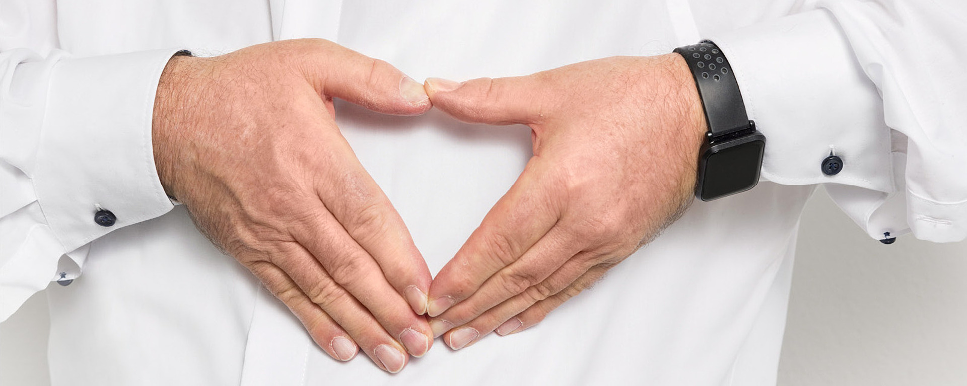 Ein älterer Mann in einem weißen Hemd lächelt und formt mit seinen Händen eine Herzform vor einem weißen Hintergrund. An seinem linken Handgelenk trägt er eine schwarze Uhr.