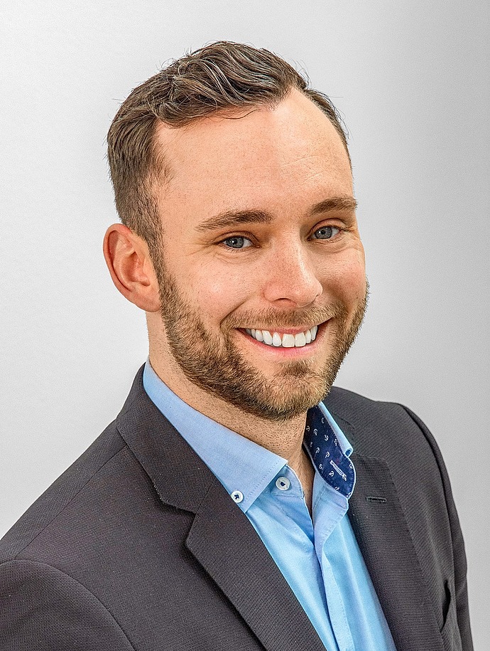 Ein lächelnder Mann mit kurzen braunen Haaren und einem Bart, der eine dunkle Anzugsjacke und ein hellblaues Hemd trägt, vor einem schlichten hellgrauen Hintergrund.