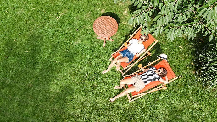 Zwei Personen entspannen sich auf orangefarbenen Liegestühlen in einem grasbewachsenen Garten unter einem Baum. Einer trägt kurze Hosen und ein gestreiftes Hemd, der andere ein weißes Hemd und eine Sonnenbrille. In der Nähe steht ein kleiner runder Tisch. Die Szene ist sonnig und friedlich.