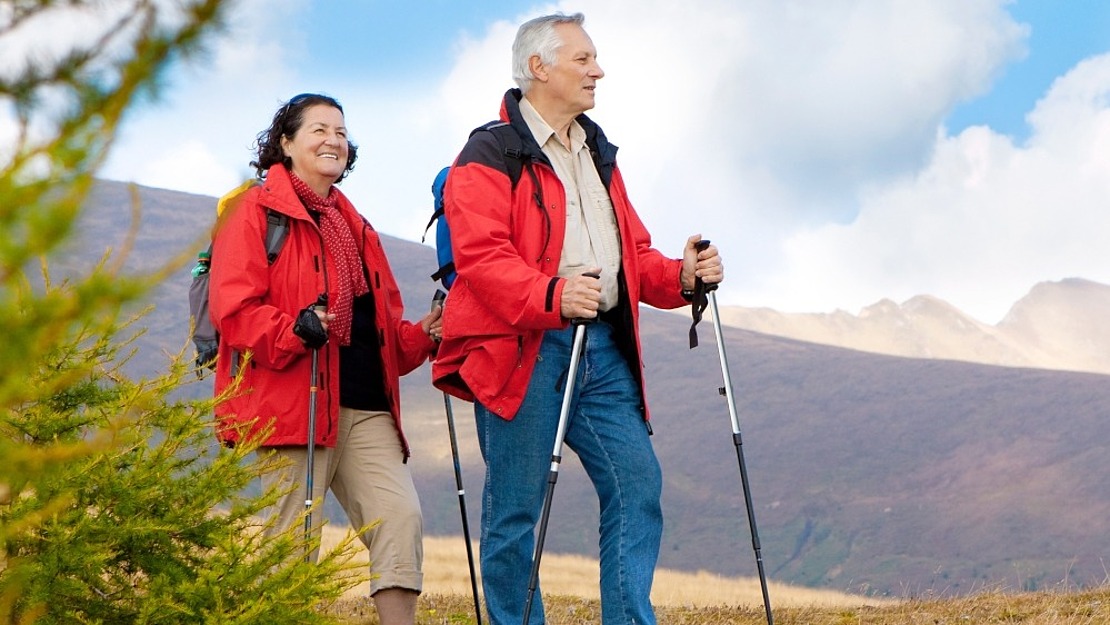 Zwei ältere Erwachsene in roten Jacken und Wanderausrüstung gehen mit Trekkingstöcken auf einem grasbewachsenen Hügel, mit Bergen und einem teilweise bewölkten Himmel im Hintergrund. Beide lächeln und scheinen die Wanderung zu genießen.