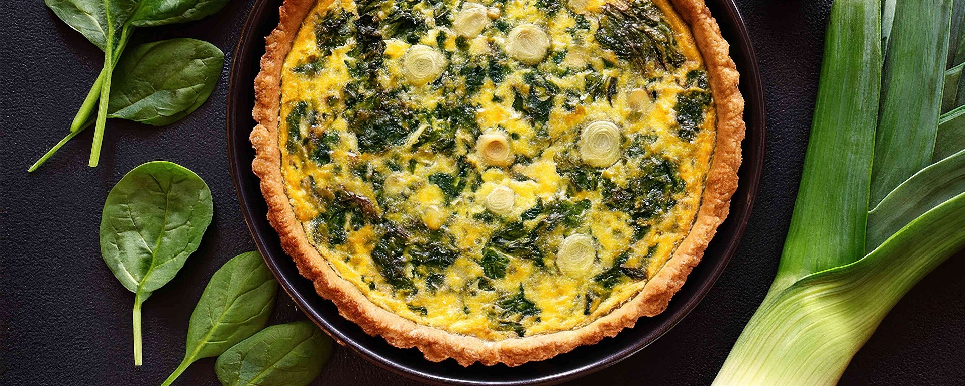 Eine Gemüsequiche in einer Kuchenform ist von frischen Spinatblättern, einer Lauchstange und zwei ganzen Champignons umgeben, alles auf einer dunklen Oberfläche präsentiert.